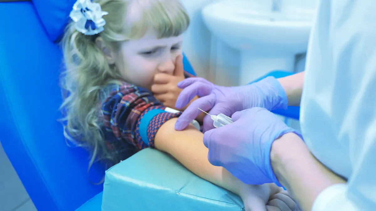Ein Kleinkind sitz in einem Stuhl und eine Kanüle ist im Vordergrund bei einem medizinischen Fachpersonal in der Hand